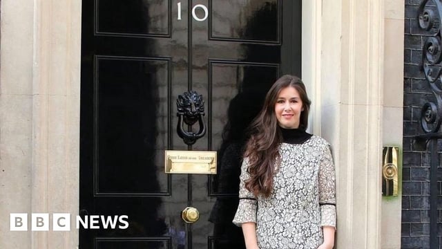 Zoe Clark-Coates is standing outside the black door of No.10 Downing Street. She has long dark hair and is smiling.