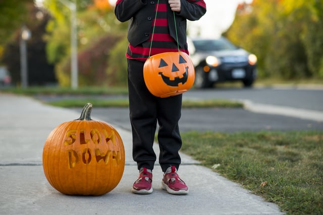 Closeup of a young boy wearing a Halloween pirate costume and holding a pumpkin bucket for trick or treat candy, standing next to a jack-o-lantern carved with the words " slow down ". This is a message encouraging vehicle drivers to practice road safety and caution so they are not speeding on the roads in urban areas. In the background a car is driving by.