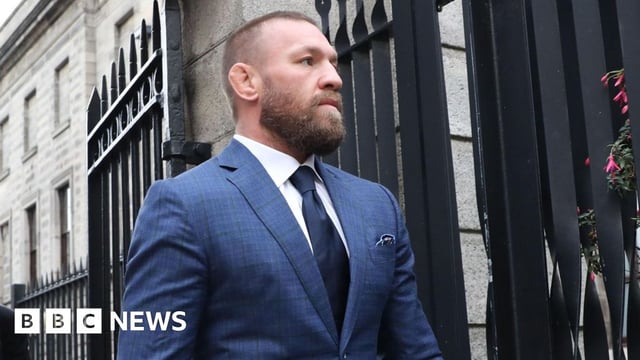 Two men walk out a black gate, one behind the other. The man at the front is wearing a navy suit, navy tie and white shirt, he has ginger short hair and a ginger beard.
The man behind him wears a black suit and tie with a light blue shirt.