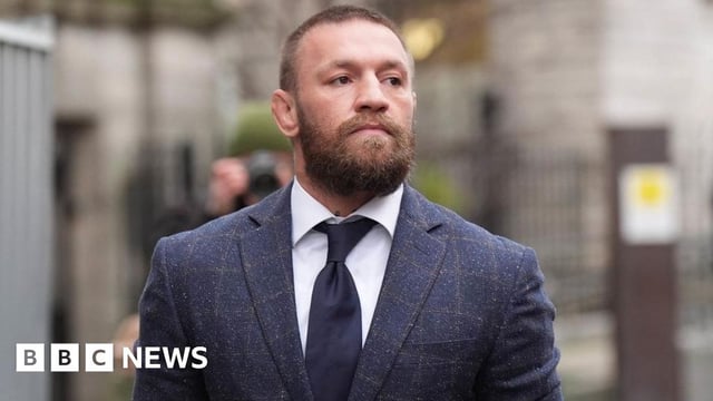 Conor McGregor walks outside court. He has cropped, light brown hair and and a beard. He is wearing a checked blue suit, light shirt and navy tie and is looking to his side.
