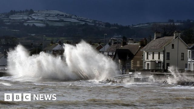 Large white waves crash against the road during a storm. There are a number of visible houses. In the background there are hills covered in snow.