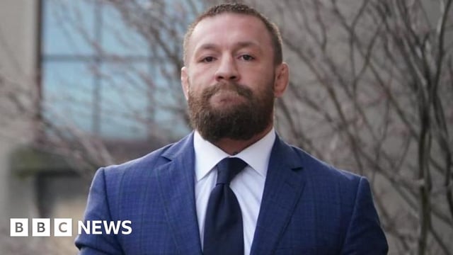 McGregor is wearing a navy suit with a white shirt and dark coloured tie. He has a thick brown beard and short brown hair. He has a straight face looking directly at the camera.