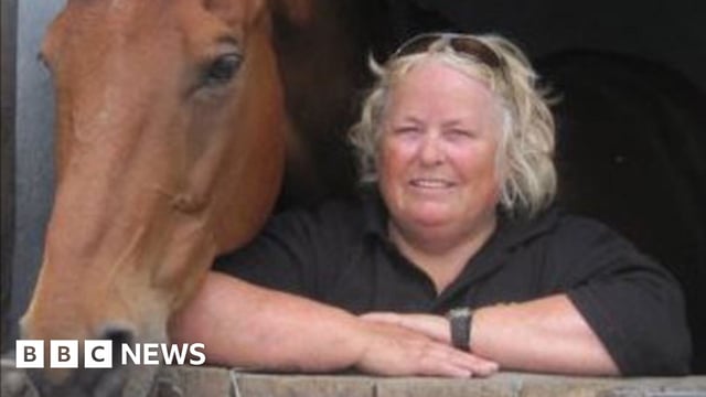 Wendy Buckney leans on a stable door, with a brown horse next to her. Wendy has blonde, shoulder length hair and is smiling at the camera. She is wearing a black T-shirt