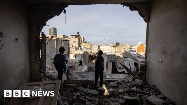 Two Palestinian children stand inside a destroyed apartment, looking out of the space where a wall used to be across a landscape of destroyed buildings