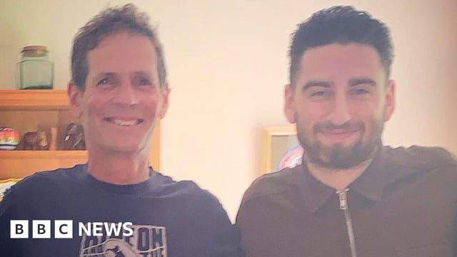 A picture of Mr Green and Mr Herries who stand arm in arm and smile at the camera. Mr Green has dark hair and a full beard while Mr Herries is clean-shaven.