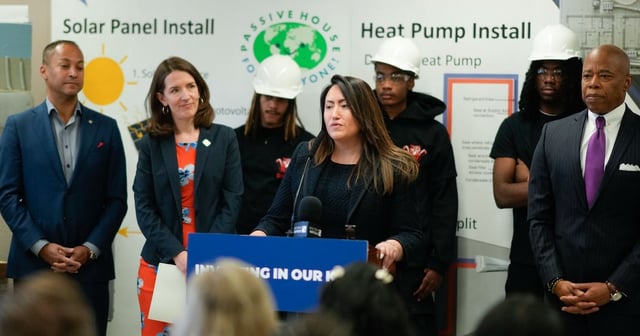 A woman in a dark suit speaks from behind a podium while a group of adults stand behind her.
