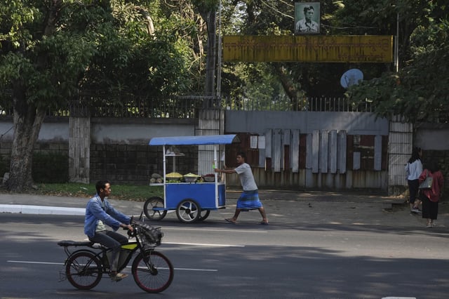A man rides bicycle while a fruit vendor pushes his cart outside the residence of ousted leader Aung San Suu Kyi as the home is auctioned in Yangon, Myanmar, Wednesday, Feb. 5, 2025. (AP Photo/Thein Zaw)