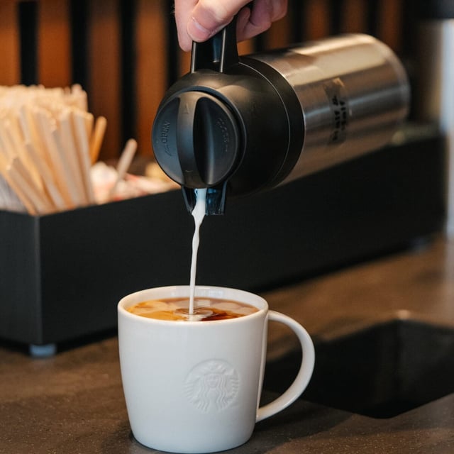 a cup of starbucks coffee in a white mug sits on a table while someone pours creamer into the coffee