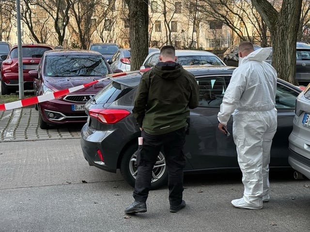 Zwei Beamte der Spurensicherung stehen vor dem Auto der Opfer, das mit Absperrband gesichert ist.