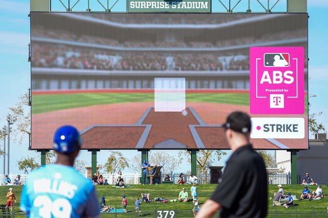 Kansas City Royals first base coach Damon Hollins and umpire Chris Guccione look on as the results of a Automated Ball Strike (ABS) replay challenge by Texas Rangers catcher Chad Wallach are shown on the scoreboard during the eighth inning of a spring training game at Surprise Stadium on Feb. 21, in Surprise, Arizona.  Wallach successfully challenged a called ball. (Smiley N. Pool/The Dallas Morning News/TNS)