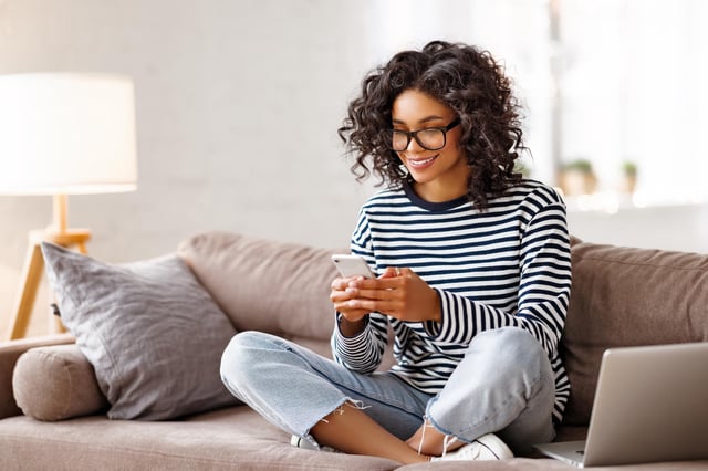 A woman wearing glasses, a striped shirt and jeans sits on a couch with her laptop and mobile phone.