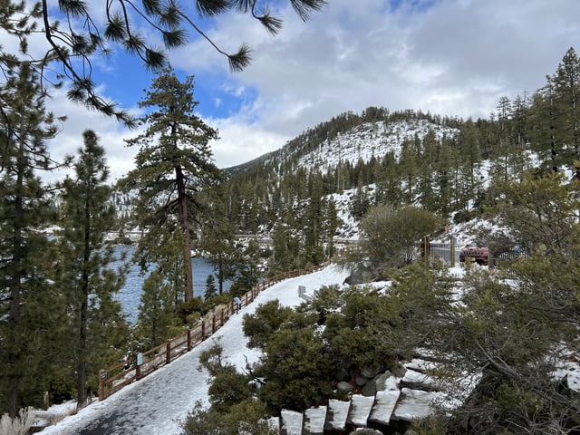 Snow-covered walkway and pine trees along the Tahoe East Shore Trail with Lake Tahoe and forested hillsides in view, Incline Village, Nevada, February 16, 2025.