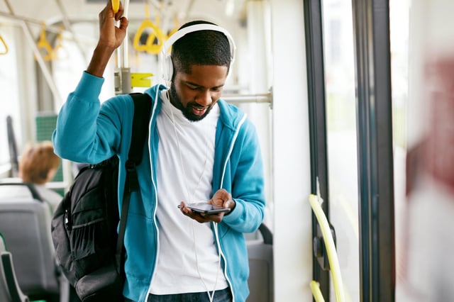 In this stock image, a man wearing a backpack and a blue sweatshirt listens to music on a train.