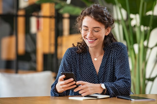 In this stock image, a woman wearing glasses and a necklace uses her mobile phone while sitting at a desk in an office.
