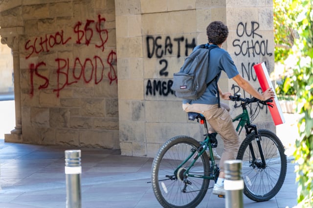 A bicyclist examines pro-Palestinian graffiti on the walls at Stanford's Main Quad on June 5, 2024. Earlier that morning, protesters barricaded themselves inside the Stanford University building that houses the office of the president and tagged multiple sites around the quad. Photo by Anna Hoch-Kenney.