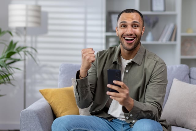 In this stock image, a man celebrates at home on the couch while holding his mobile phone.