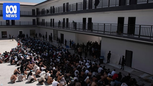 Men sit outside apartment building in rows in the sun.
