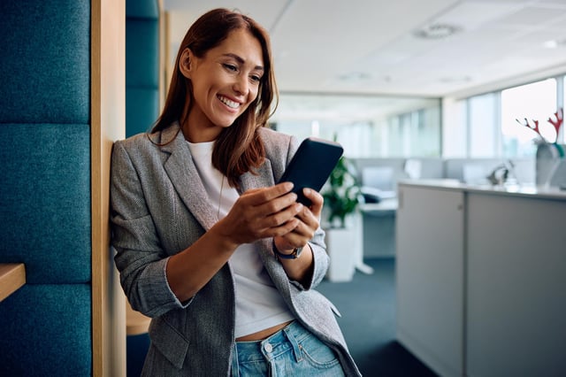 In this stock image, a woman smiles while sending a text message on her mobile phone in an office.