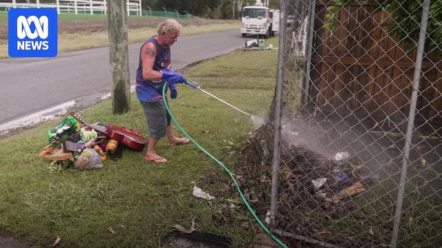 A man hosing down his property, debris can be seen on the ground nearby.