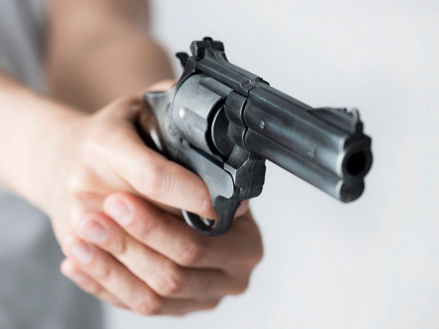 Unrecognizable person wearing a grey t-shirt is holding a gun with two hands in front of grey background.