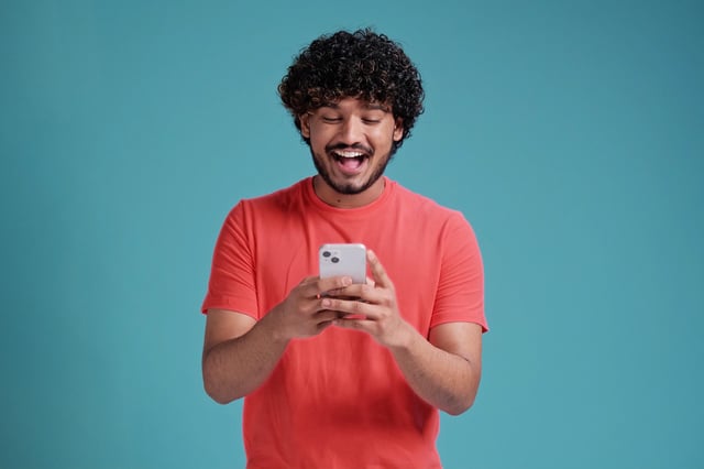 In this stock image, a man wearing a coral shirt against a blue background smiles while looking at his mobile phone.