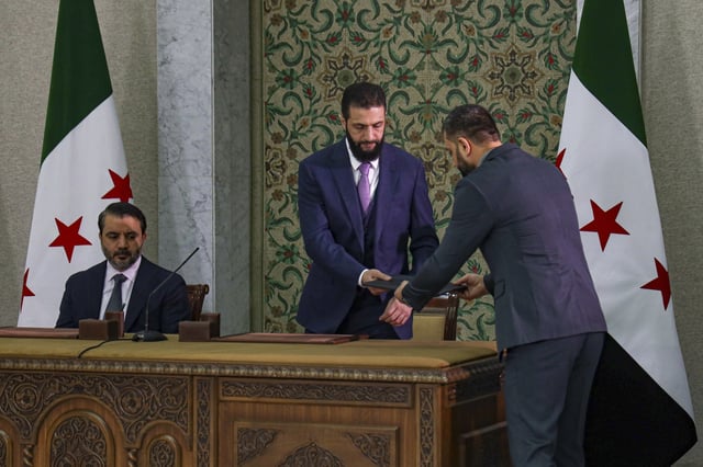 Syria's interim president Ahmad Al-Sharaa, center, prepares to sign a temporary constitution for the country in Damascus, Syria, Thursday March 13, 2025. At left, is Foreign Minister Asaad Hassan al-Shiban. (AP Photo/Omar Albam)
