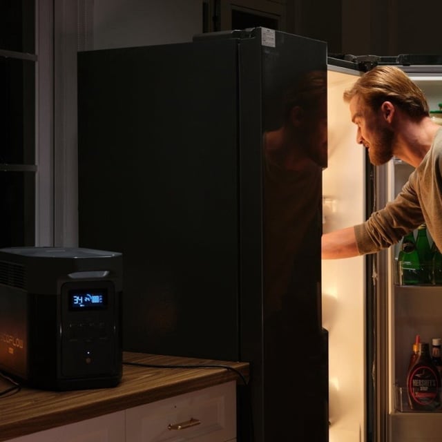 the EcoFlow Delta 2 Max portable power station sits on a kitchen countertop providing power for a refrigerator while a person is standing in front of the open fridge removing some food