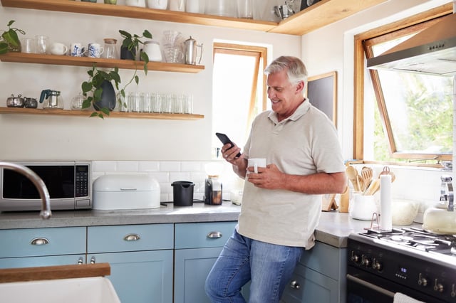 In this stock image, a man wearing blue jeans smiles in his modern kitchen while holding a mobile phone and a cup of coffee.