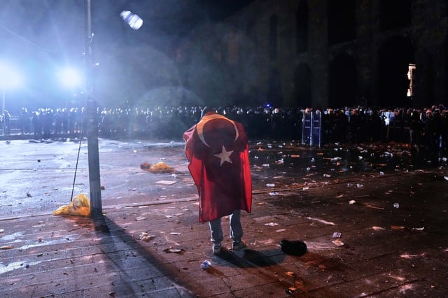A man, a Turkish flag draped on his back, stands in front of anti riot police officers during clashes in a rally against the arrest of Istanbul's Mayor Ekrem Imamoglu, in Istanbul, Turkey, Friday, March 21, 2025. (AP Photo/Khalil Hamra)