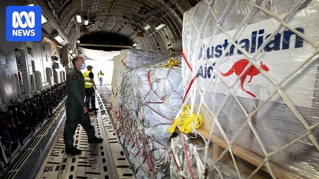 A man in military uniform inside an aircraft stand in front of palettes carrying items labelled 'Australian Aid'.