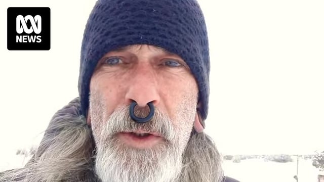 A man with a septum piercing and white beard and long hair wearing a beanie outside while it snows