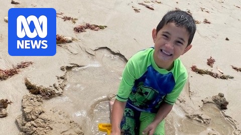 Caleb sitting on the beach digging a hole in the sand smiling.