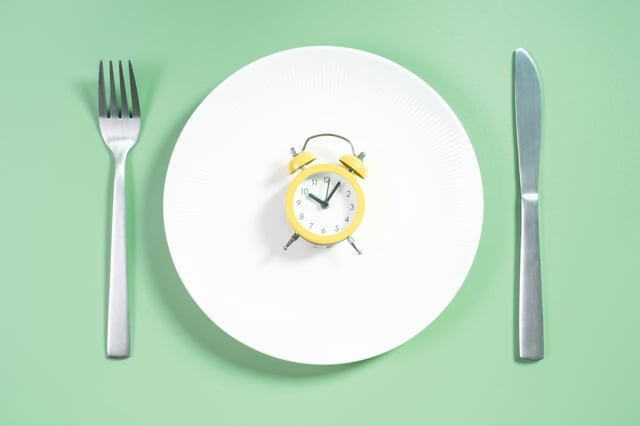 Empty plate with alarm clock on green background, intermittent fasting concept.