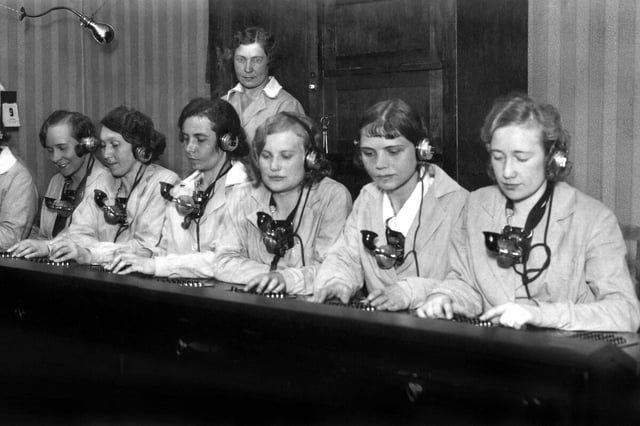 Telefonistinnen sitzen mit Kopfhörer an einem Tisch mit Schalthebeln - schwarz-weiss Aufnahme aus den 30er Jahren