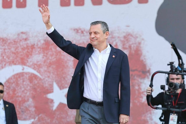 Özgur Özel steht auf einem Podiumund hebt grüßend die Hand. Er trägt ein weisses Hemd, blaues Jacket und einen CHP-Sticker am Revers