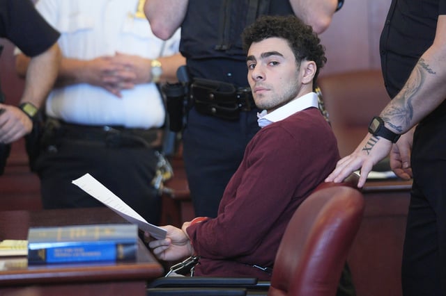 A man with dark brown curly hair, thick eyebrows and a five o'clock shadow looks into the camera, sitting in front of a courtroom desk. He's wearing a dark maroon sweater with a white collar underneath and holding a piece of paper. Police stand beside him. 