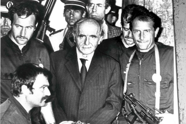 Klaus Barbie, chef de la Gestapo de Lyon pendant la Seconde Guerre mondiale, est photographié menotté alors qu'il est conduit hors de la salle d'audience par des policiers lourdement armés après avoir été condamné à la prison à vie le 4 juillet 1987 à Lyon, en France, pour crimes contre l'humanité.
