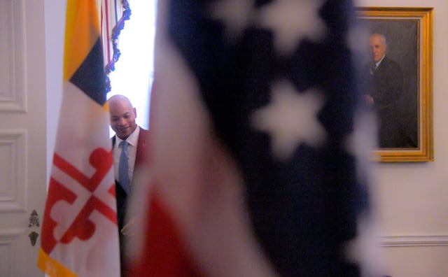 Framed between the flags of Maryland and the United States, Wes Moore, D-Md. smiles while entering the conference room filled with paintings of past gubernatorial leaders including Gov. Marvin Mandel to speak with the media during the final day of the 2025 legislative session at the Maryland State House. (Karl Merton Ferron/Staff)