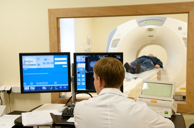 A doctor looks at two computer screens outside a CT scan room where a patient is moving through the scanning machine -- Health coverage from STAT
