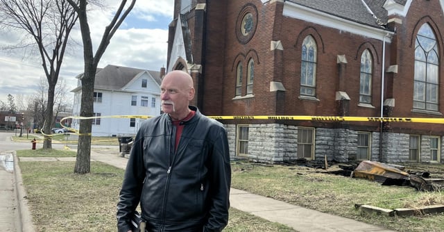 A man in a leather jacket stands in front of a brick church surrounded by yellow caution tape.