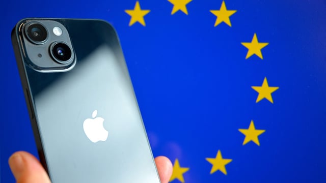 This photograph shows a hand-held smartphone with the Apple company logo and the European Union logo in the background,