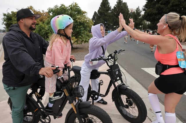 John Hodge and daughter Tatum, 5, watch as his wife Vineca Hodge high fives daughter Marlee, 13, who are following on e-bikes, as she and thousands of people participating in the half marathon run on Harbor Island during the 47th annual America’s Finest City Half Marathon and 5K in San Diego on Sunday, Aug. 18, 2024. Over 1,300 people participated in the 5K and more than 4,300 in the half marathon. (Hayne Palmour IV / For The San Diego Union-Tribune)