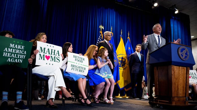 Robert F. Kennedy Jr. speaking at a podium next to a group of MAHA moms 