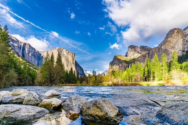 Landscape image of El Capitan and Half Dome in Yosemite National Park with the rocky shore of the Merced River in the foreground.