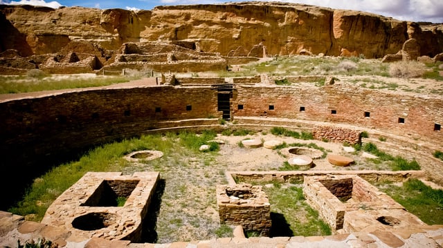 Image of a large, circular, below-ground area surrounded by very finely crafted stone walls. Other ruins are in the background.