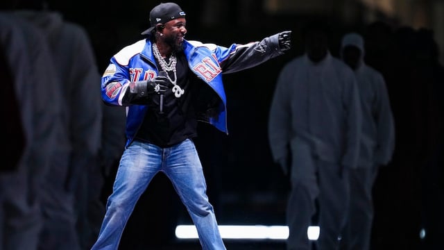 Kendrick Lamar smiles and points as he performs in a blue and black leather jacket, blue jeans, gloves and a black backwards cap