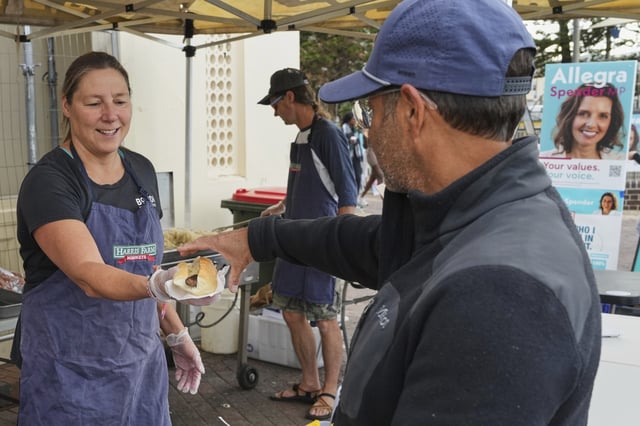 A volunteer hands a customer a sausage in a bread roll outside a polling booth at Sydney's Bondi Beach, Saturday, May 3, 2025. (AP Photo/Mark Baker)