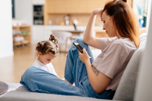 Mom using smartphone at home on couch while with her toddler.