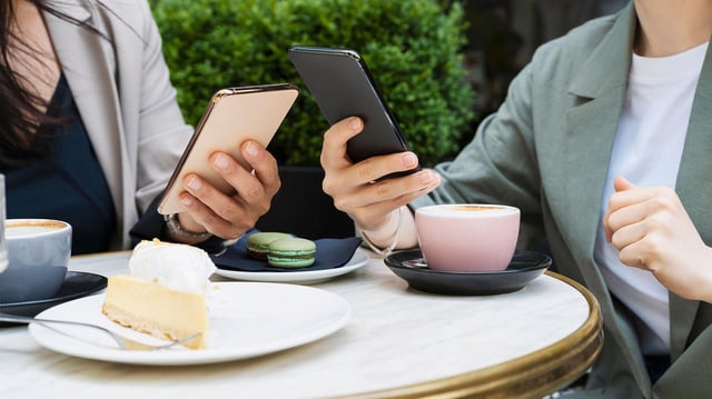 Zwei Personen nutzen Smartphones bei Kaffee und Kuchen auf einem Tisch im Straßencafé.