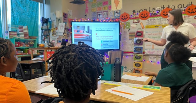 A woman in white points to a screen as students look on.